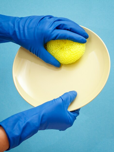 Women's hands in protective gloves with beige plate and yellow sponge on blue background. Washing and cleaning concept.