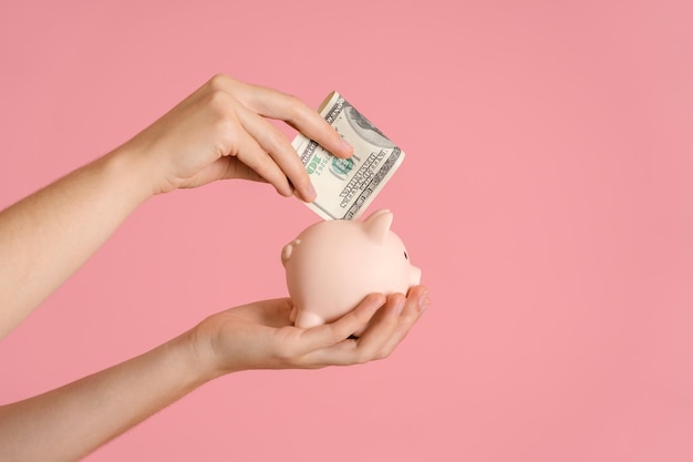 Women's hands put a hundred-dollar bill folded in half in a piggy bank, pink background copyspace