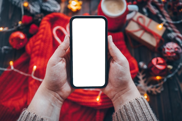 Women's hands in a warm knitted sweater with a smartphone holder with an isolated screen on Christmas background with gifts Happy New Year and Merry Christmas