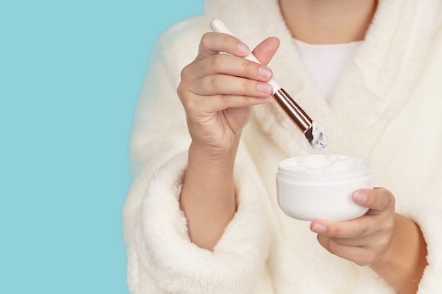 Women's hands with a jar of face cream and a cosmetic brush on an isolated blue