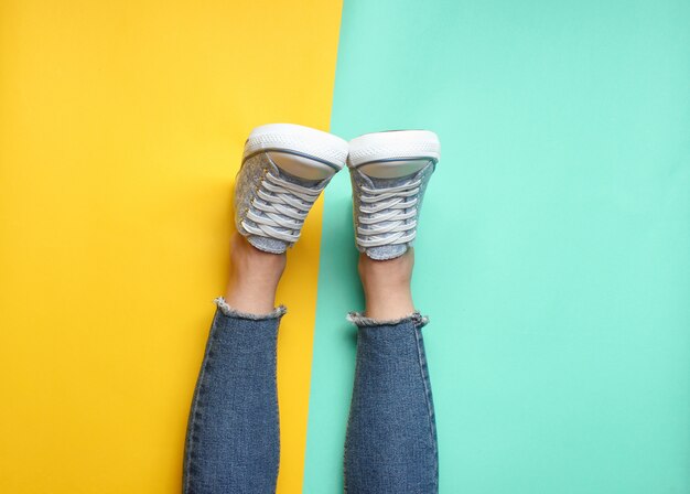 Women's legs in tight, torn jeans, sneakers on yellow blue pastel. Top view