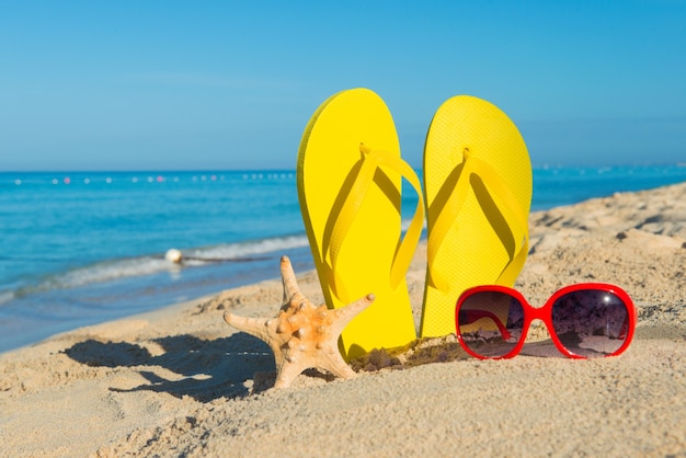 Women's red sunglasses and yellow flip-flops on sandy seashore. Travel by sea. Beach vacation