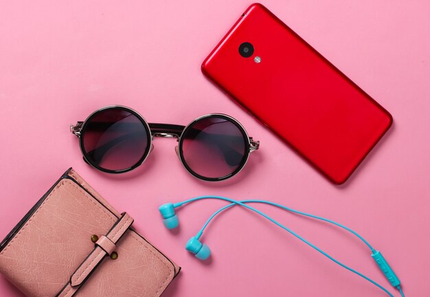 Womens accessories and gadgets on a yellow. Earphones, stylish round sunglasses, smartphone, wallet on pink