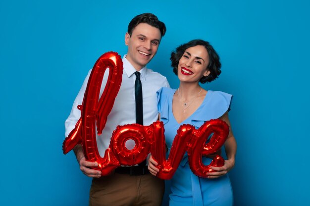 Womens Day Valentines Day Birthday Holidays presents Excited handsome young couple in love celebrating Valentines Day with red love shaped balloons isolated over blue background