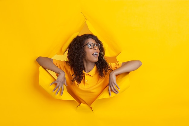 Wondered happy. Cheerful african-american young woman in torn yellow paper background, emotional and expressive. Breaking on, breakthrought. Concept of human emotions, facial expression, sales, ad.