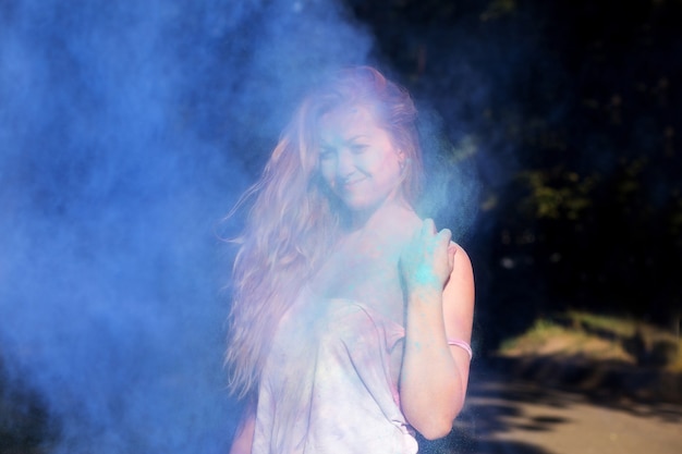 Wonderful blonde woman with curly hair posing in a cloud of blue powder at Holi festival of colors