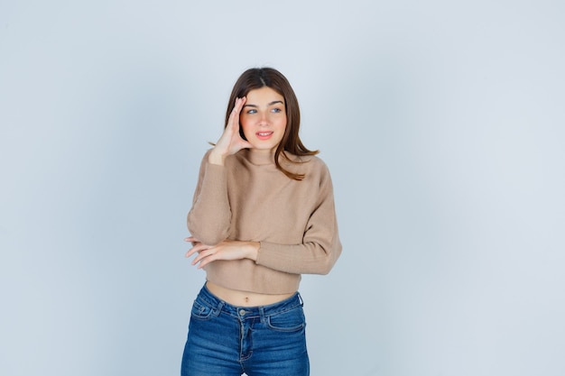 Wonderful lady keeping hand on head, looking aside in sweater, jeans and looking interested , front view.