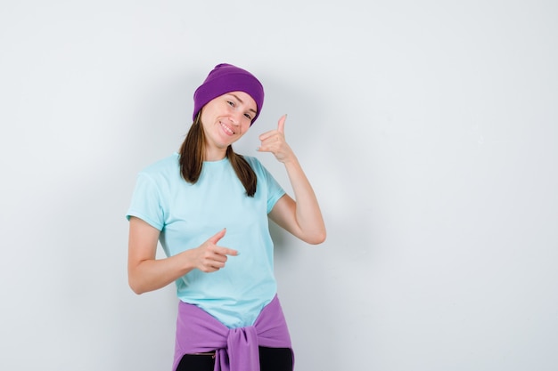 Wonderful lady showing phone gesture in blouse, beanie and looking merry , front view.