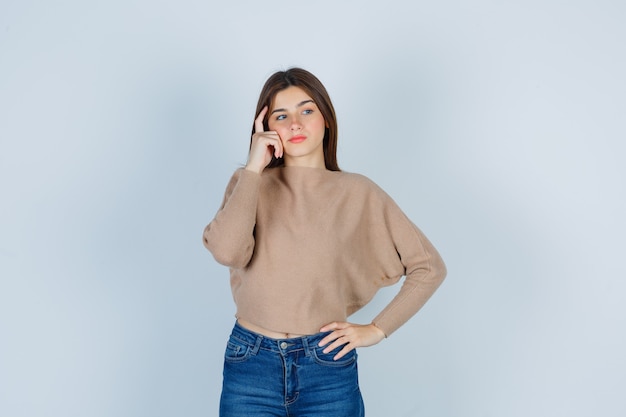 Wonderful lady in sweater, jeans keeping hand on cheek while looking aside and looking pensive , front view.