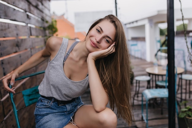 Wonderful longhaired female model sitting on chair in street cafe Romantic white girl in denim skirt resting in outdoor restaurant in summer morning