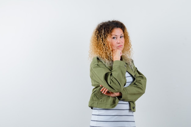 Wonderful woman in green jacket, shirt keeping hand on chin and looking preoccupied , front view.