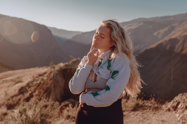 A wonderful young woman stands under the bright hot sun in the rocky mountains with her eyes closed