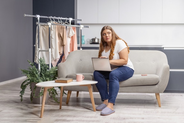 Wondering looking at the window young fat girl working on laptop sitting on the sofa at home with the kitchen