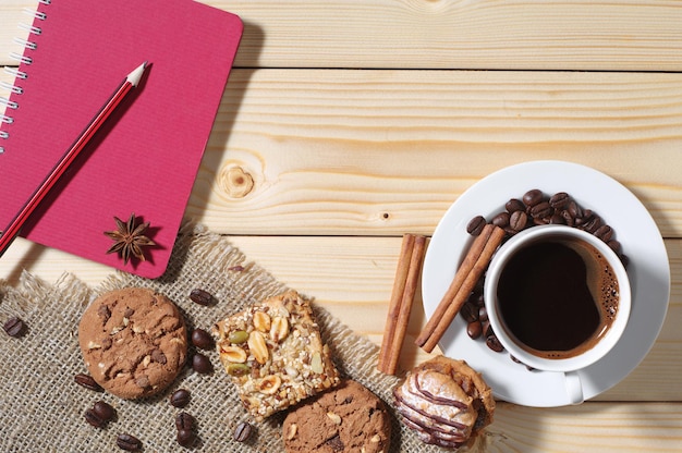 Wooden background with cup of hot coffee, different cookies and notepad. Top view