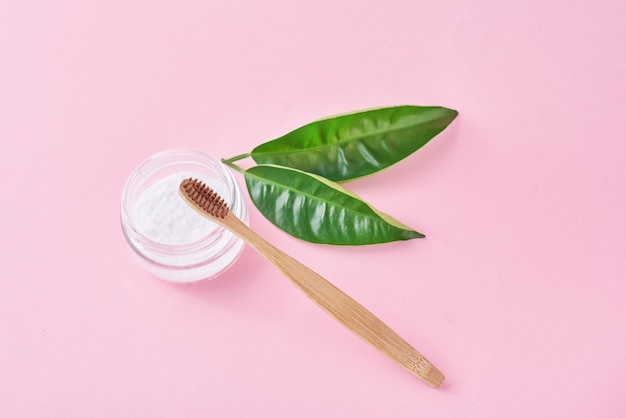 Wooden bamboo toothbrush with baking soda powder in glass jar and green leaves on a pink background. 