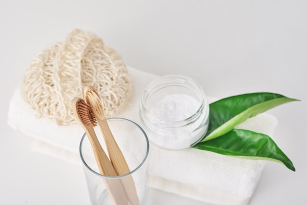 Wooden bamboo toothbrushes in glass, baking soda and towel on white.