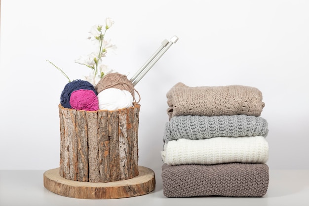 Wooden basket with balls of wool and knitting needles with wool sweaters folded on a white table and white background