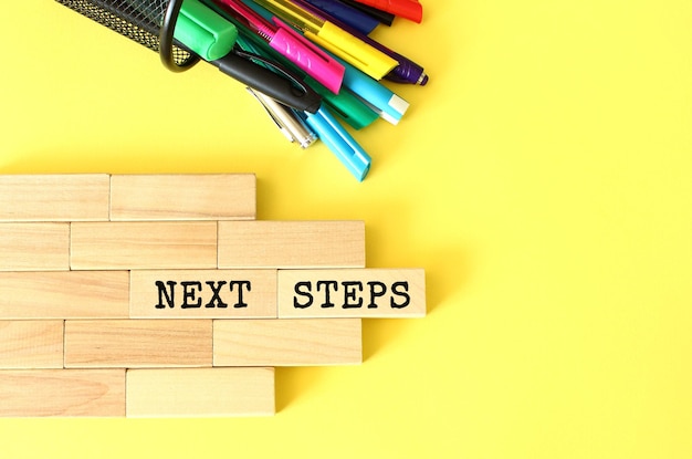 Wooden blocks stacked next to pens and pencils on a yellow background NEXT STEPS text on a wooden block