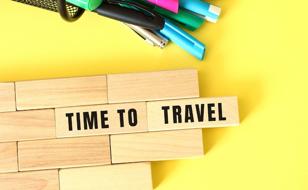 Wooden blocks stacked next to pens and pencils on a yellow background. TIME TO TRAVEL text on a wooden block.