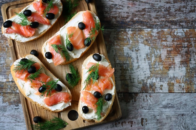 A wooden board with bread, smoked salmon, and black olives on it.