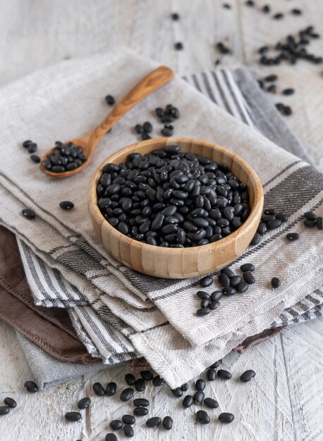 Wooden bowl full of dried black beans with a wooden spoon on kitchen towels on grey table close up. Healthy eating and vegetarian concept. Traditional Latin American cousin ingredient