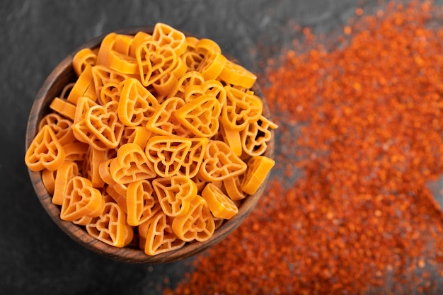 Wooden bowl of uncooked heart-shaped pasta and ground chili on black. 