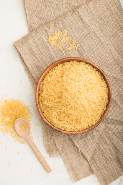 Wooden bowl with raw golden rice and wooden spoon on a white wooden table and linen textile. Top view, flat lay, close up.