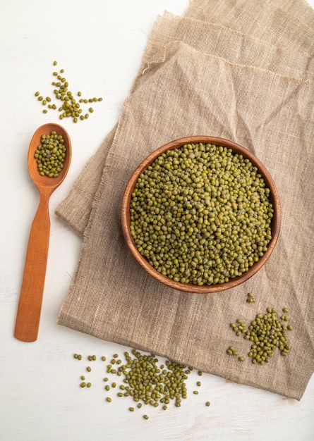 Wooden bowl with raw green mung bean and wooden spoon on a white  background and linen textile. Top view, flat lay, close up.