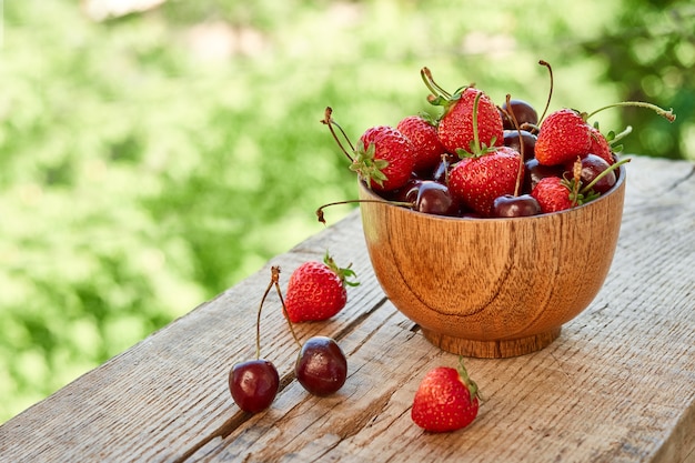 Wooden bowl with ripe red cherry, strawberry on a green background of foliage. Summer sunny still life with ripe berries