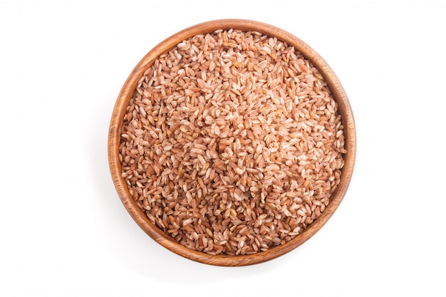 Wooden bowl with unpolished brown rice isolated on white surface. Top view, close up.