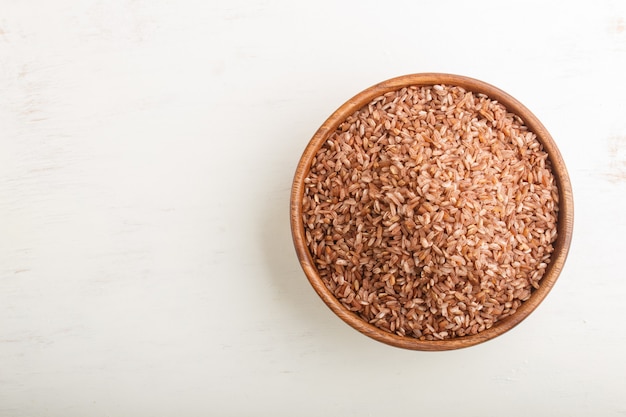 Wooden bowl with unpolished brown rice on a white wooden background. Top view, copy space.