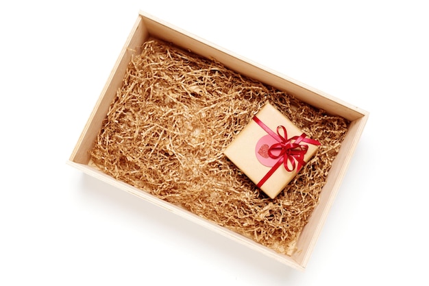 Wooden box with present wrapped in brown craft paper with red ribbon, filled with paper filler, top view from above on white background