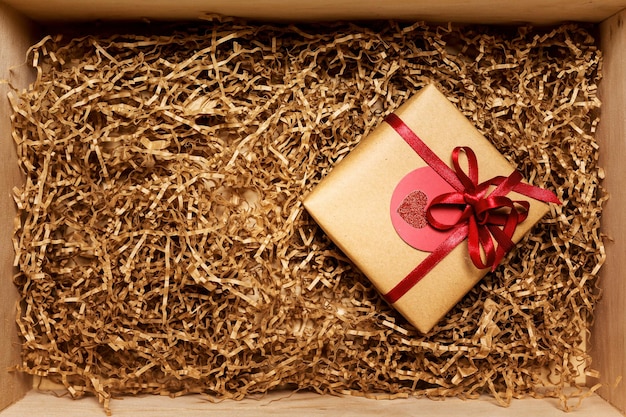 Wooden box with present wrapped in brown craft paper with red ribbon, filled with paper filler, top view from above