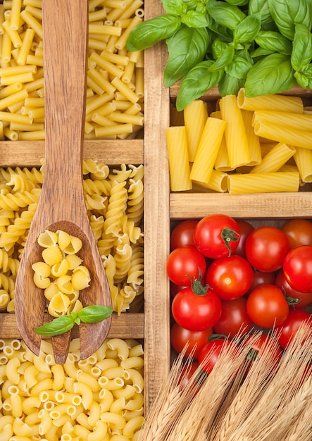 Wooden box with various classic italian pasta and basil with cherry tomatoes and wooden spoon Top view