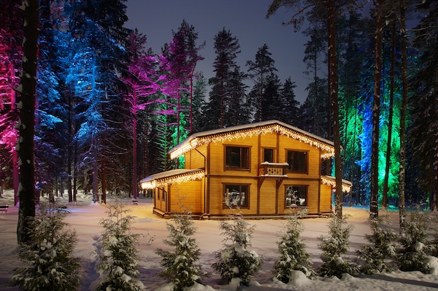 Wooden cottage with Christmas decorations in a snowy winter. Trees with bright lights in the forest near the house.