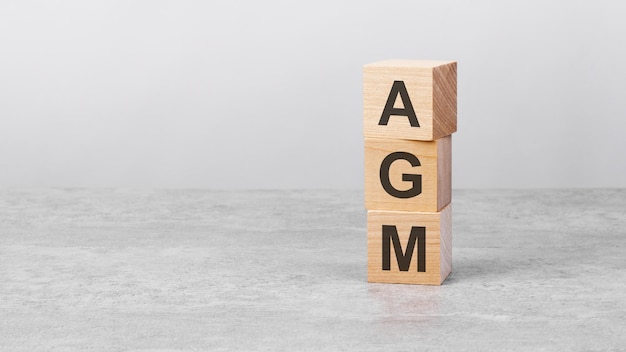 Wooden cube on a white table with text AGM concept of business financial investment economy copy space on left for you design gray background AGM  short for Annual General Meeting