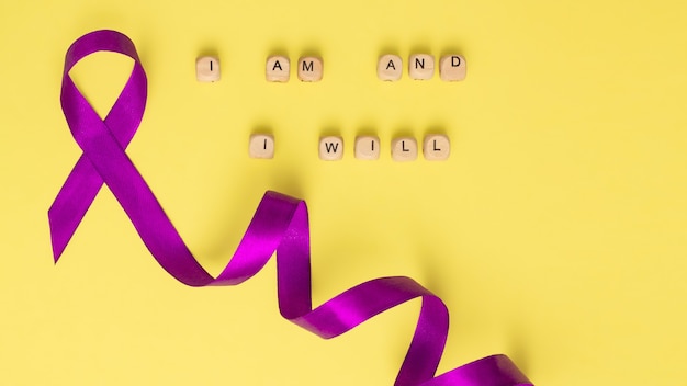 Wooden cubes with text of the slogan of World Cancer Day I am and I will and a purple ribbon, on a yellow surface. Flat lay.