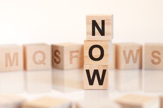 Wooden cubes with word NOW arranged in a vertical pyramid, on the white surface is a row of wooden cubes with letters