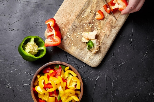 Wooden cutting board and bowl with pieces of different bell peppers on black background top view