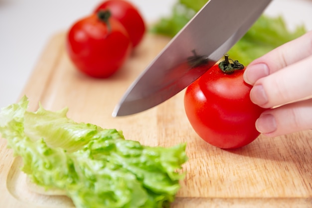 Wooden Cutting Board with Fresh Herbs, Lettuce and Raw Vegetables on Rustic Wood Board. Cooking background. Female Hands cut with a knife Juicy red tomato.