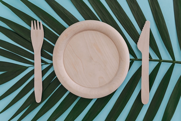 Wooden empty plate, fork and knife on palm leaf on blue. Zero waste concept.