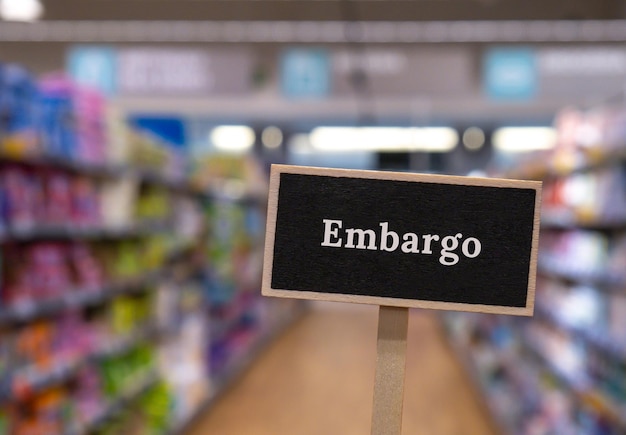 Wooden information label sign with text EMBARGO against defocused store shelves message