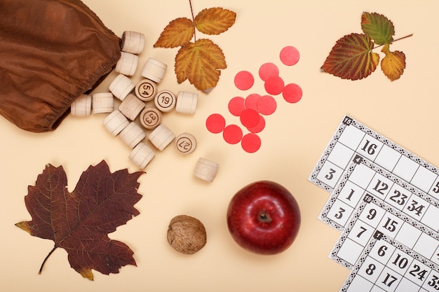 Wooden lotto barrels with open bag, dry autumn leaf, apple, walnut,, red chips and game cards on beige background. Board game lotto. Autumn theme. Top view.