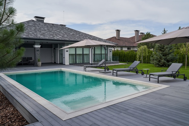 Wooden lounge chairs in modern villa pool and deck. Light blue water swimming pool of a luxurious mansion with chairs around it. Modern house at the background