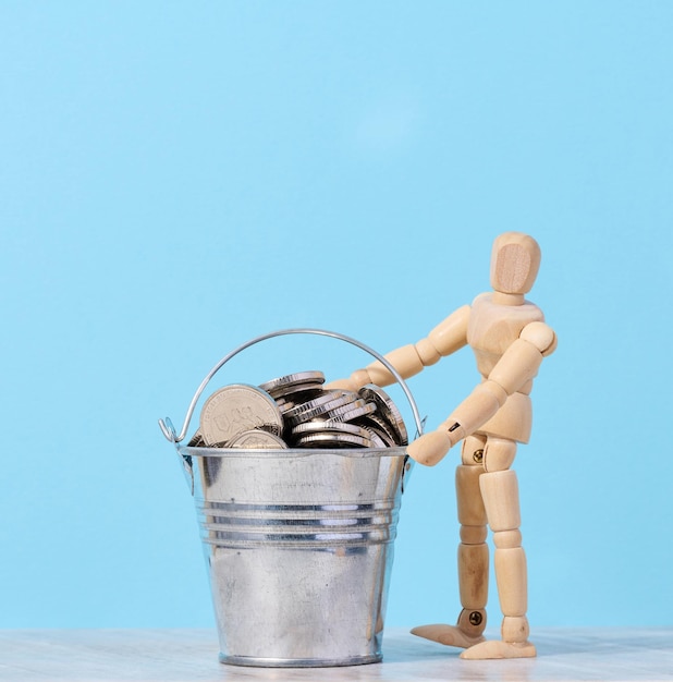 A wooden mannequin and coins in a miniature bucket on a blue background a concept of high income subsidy