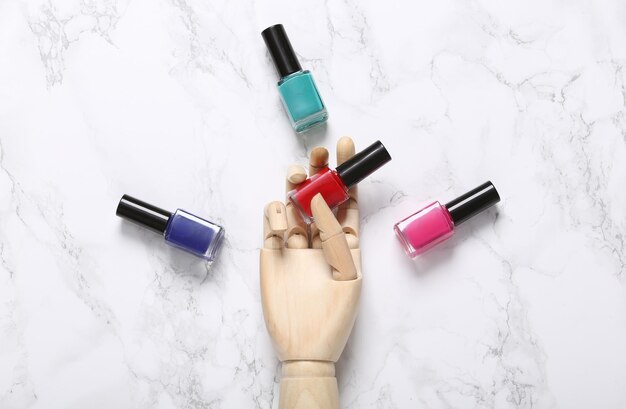 Wooden Mannequin hand holds bottles of nail polish on marble background Beauty layout