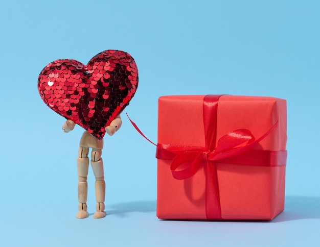 A wooden mannequin holds a red heart made of sequins and a box wrapped in red paper congratulations on the holiday