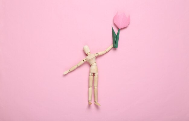 Wooden puppet hold origami tulip on pink background Top view