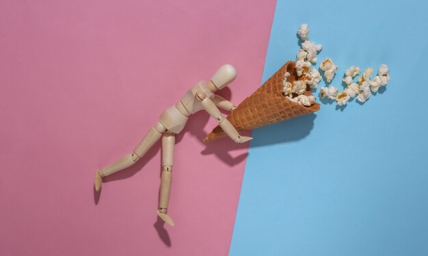 Wooden puppet holding Ice cream waffle cone with popcorn on pink blue background. Top view