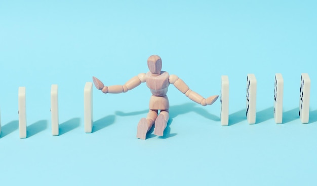 A wooden puppet toy holds back a falling domino on a blue background representing the concept of a strong personality